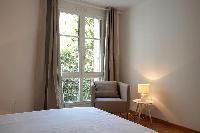 an armchair, table lamp, and French window dressed in taupe drape curtain in a 2-bedroom Paris luxur