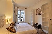 chic bedroom with a queen-size bed, shelves, chest, bedside tables, lamps, drape curtain and tall wi