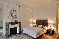 bedroom two with queen size bed, bdside tables, lamps, and ornamnetal fireplace in a Paris luxury ap