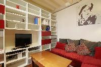 warm living area with TV, shelves, wooden center table, and an L-shaped sofa in a 1-bedroom Paris lu